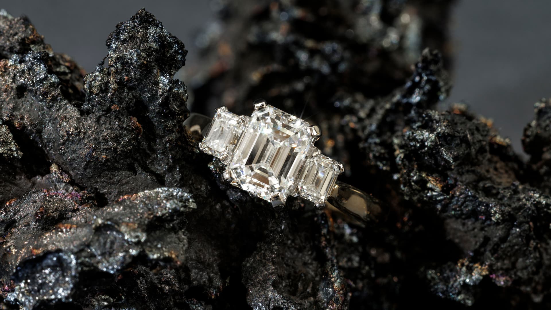 Emerald-cut three-stone diamond engagement ring from Spencer Morgan Jewellers, photographed on dark anthracite coral