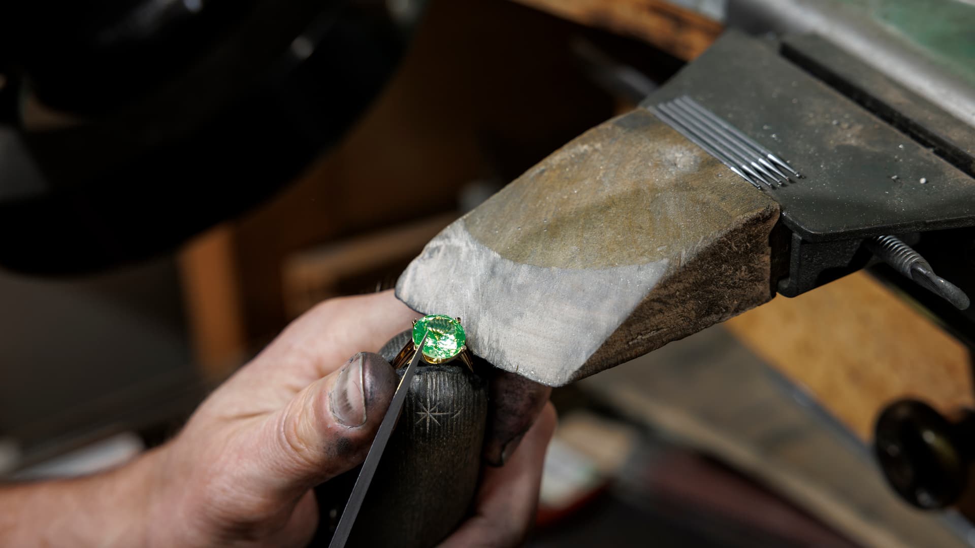 Hands at the Spencer Morgan workshop bench, setting an oval emerald in a yellow-gold claw setting, ring clamp held over the bench peg